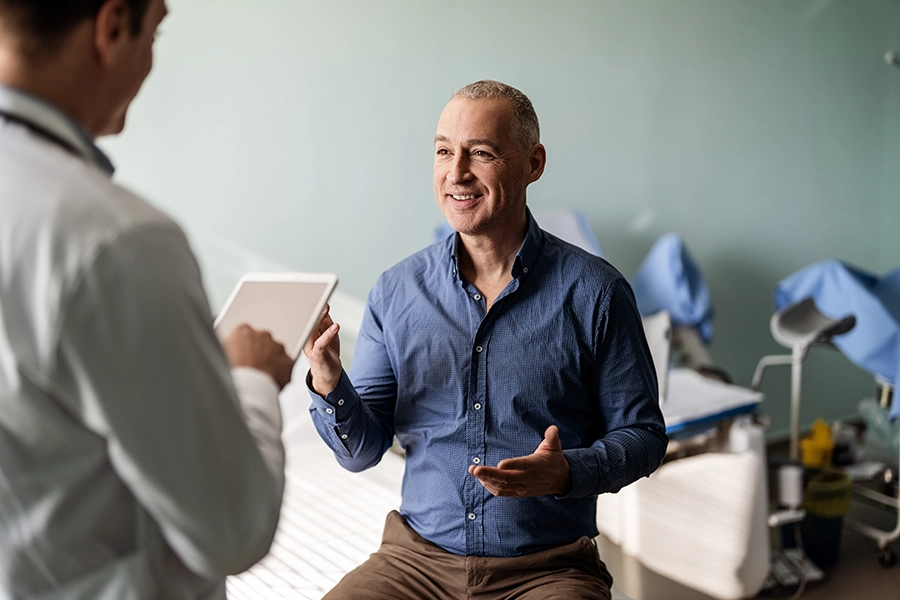 Smiling middle-aged man in a blue shirt sits on an exam table and talks with a doctor holding a tablet during a medical consultation.